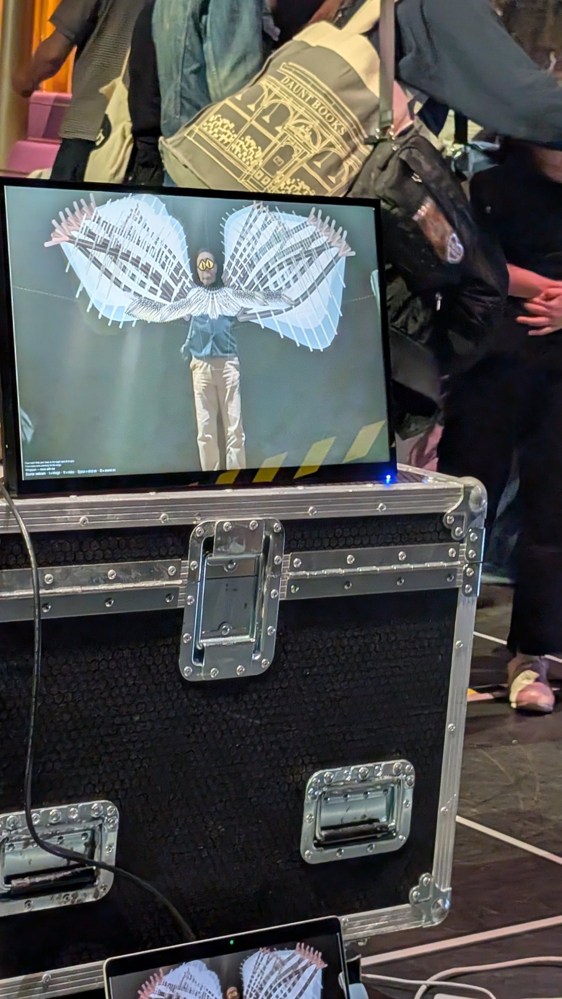 A monitor on a flight case shows a standing figure with large digital wings.