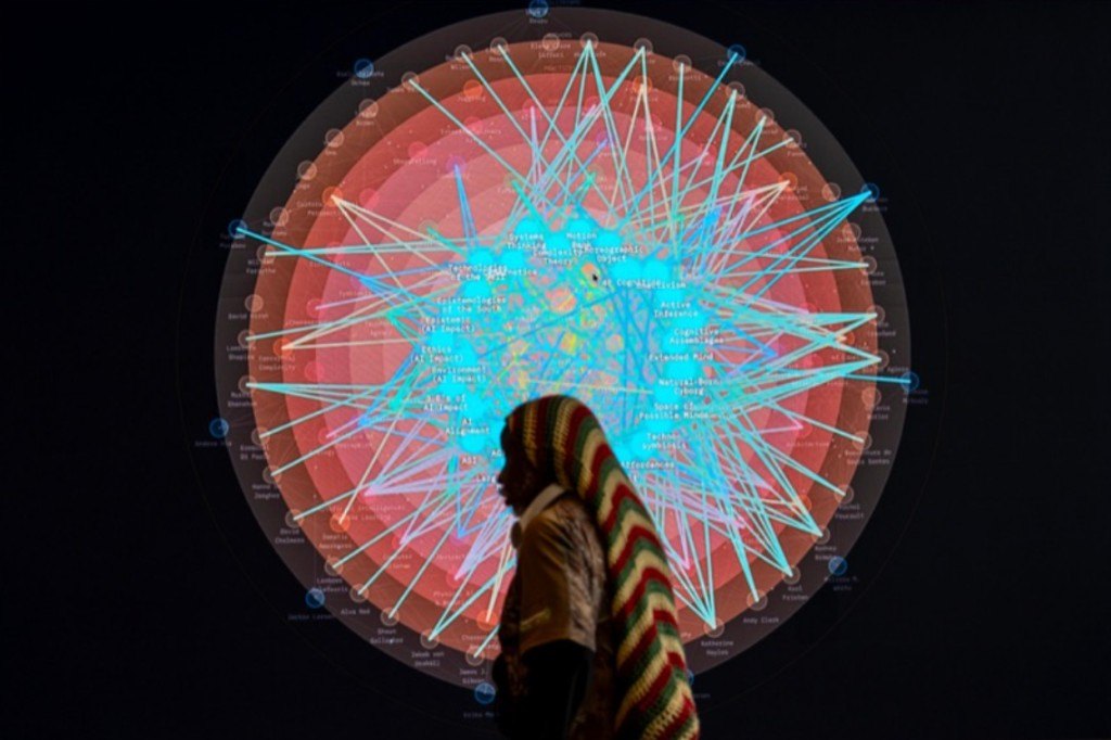 A person standing in profile before a large circular Hybrid Intelligences network visualization: concentric rings of concepts, authors, and relations glowing from a cyan core.