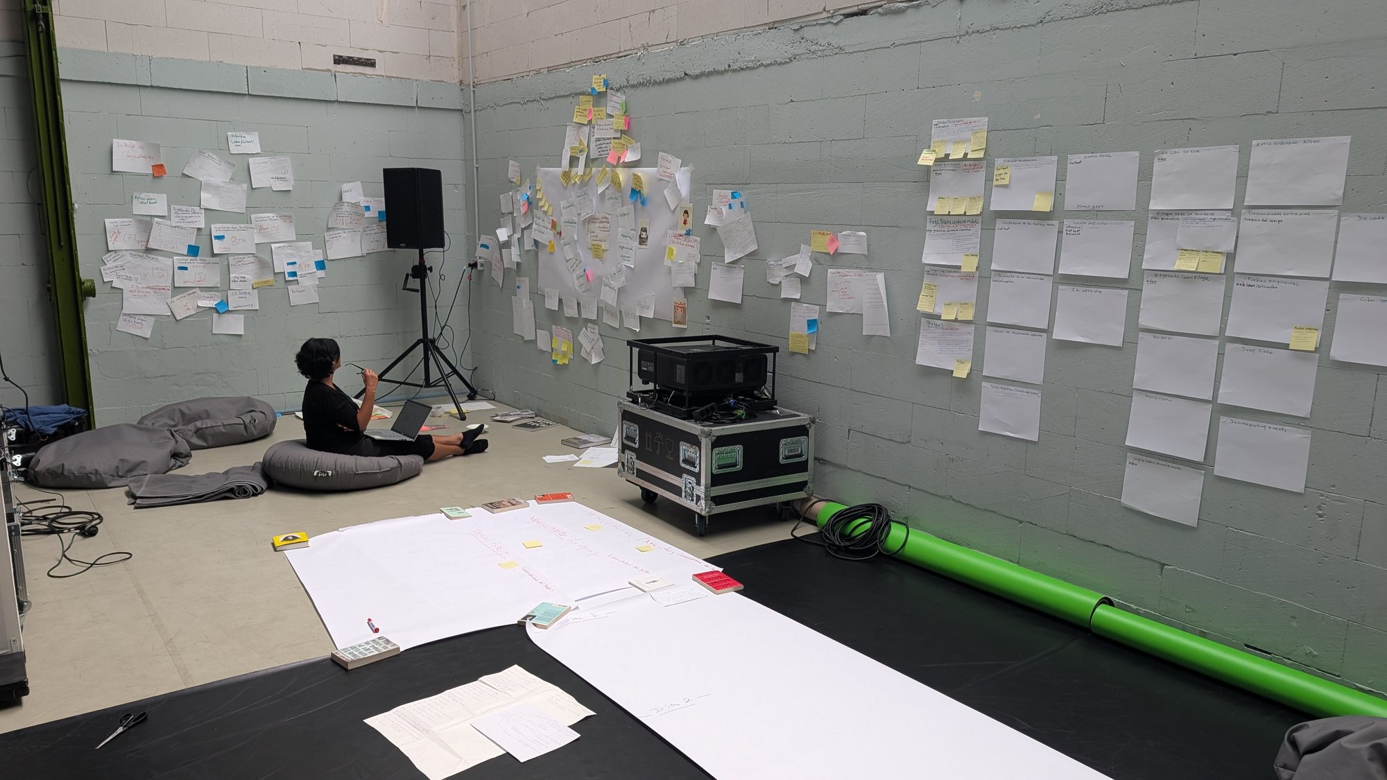 A person works on a laptop in the studio, with notes covering the walls and a large sheet of paper on the floor.