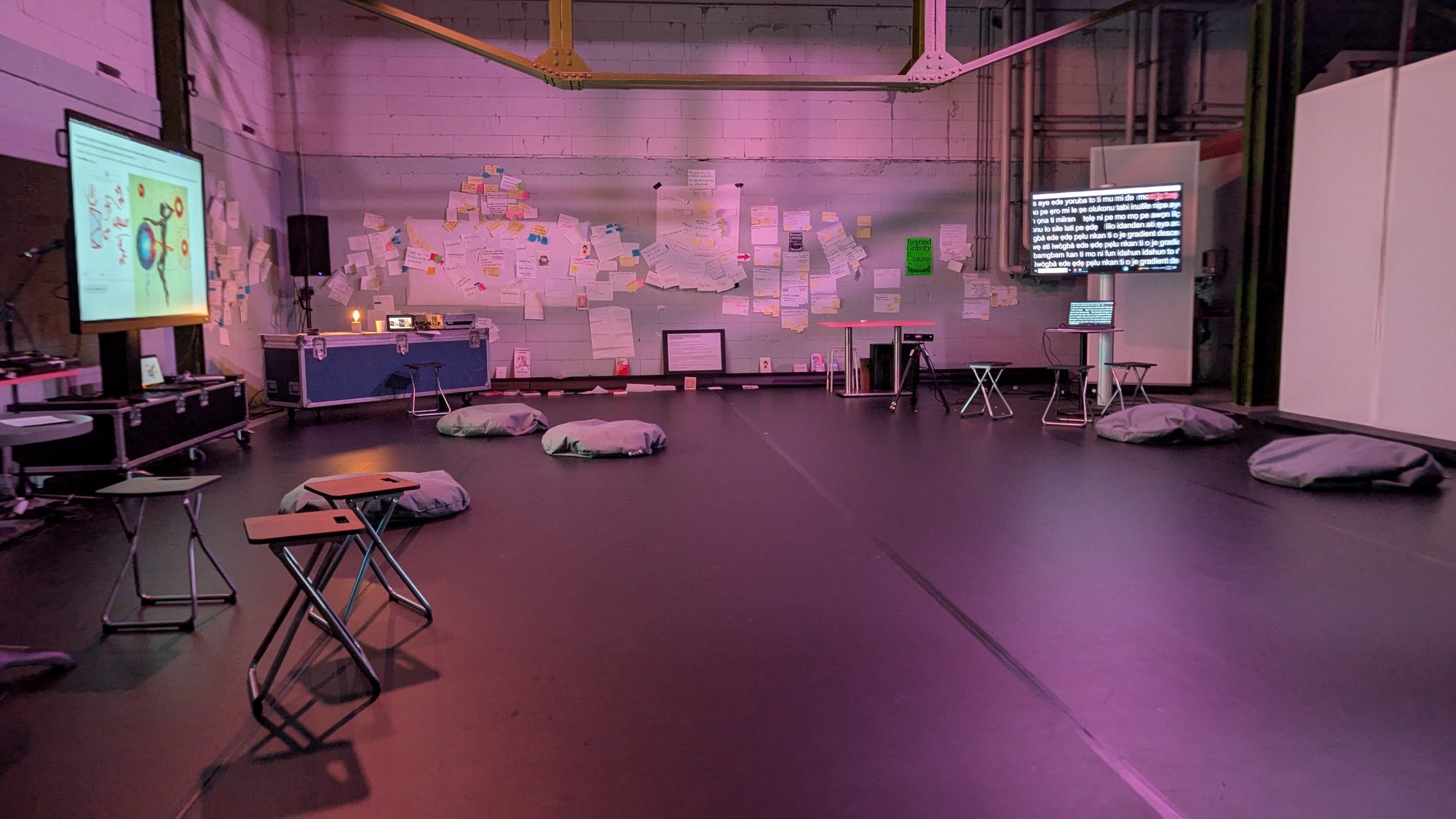 An empty studio in magenta light, with cushions, screens of text, and a wall of notes, at Beyond Gravity Festival, 2025.